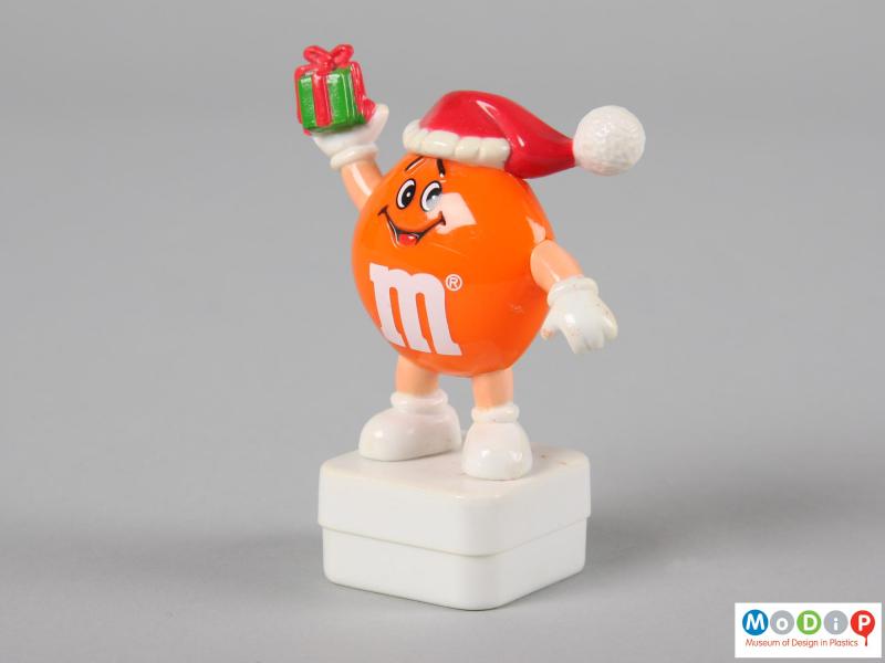 Side view of an orange M&M figure showing on hand held down and the other in the air.