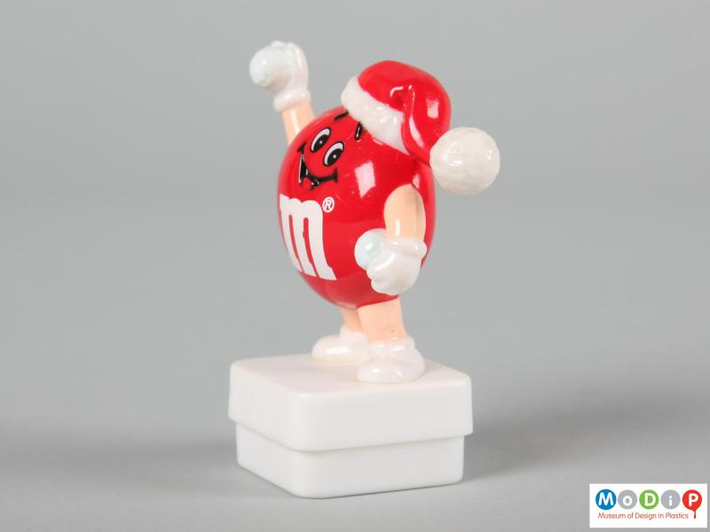 Side view of a red M&M figure showing on hand held down and the other in the air.