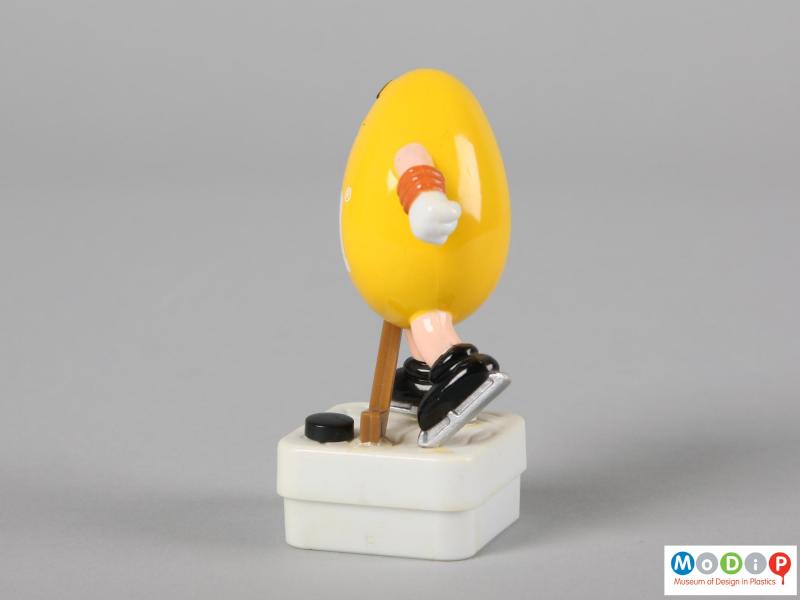 Side view of a yellow M&M figure showing one hand holding the hockey stick and the held behind.