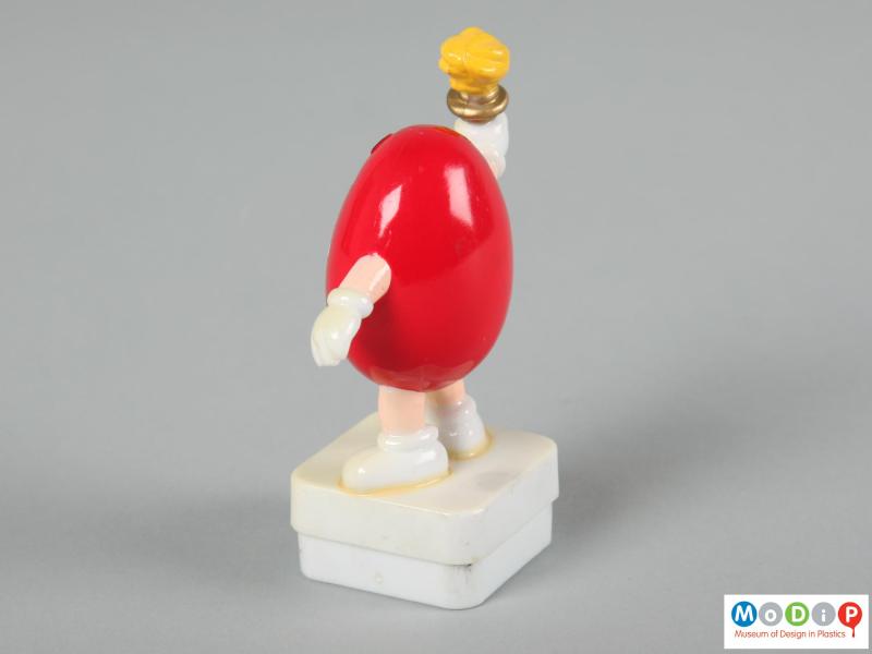 Side view of a red M&M figure showing one hand the torch aloft the other held down low.