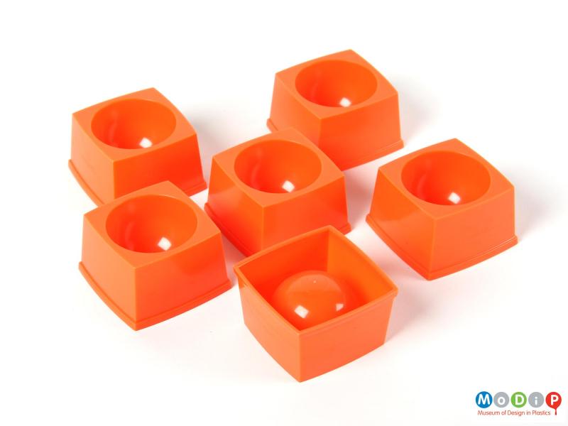 Top view of six stackable egg cups showing the square outer edghe and the half shpericla indentation for the egg to sit in.   One cup is upside down exposing the underside of the cup including the injection moulding gate.