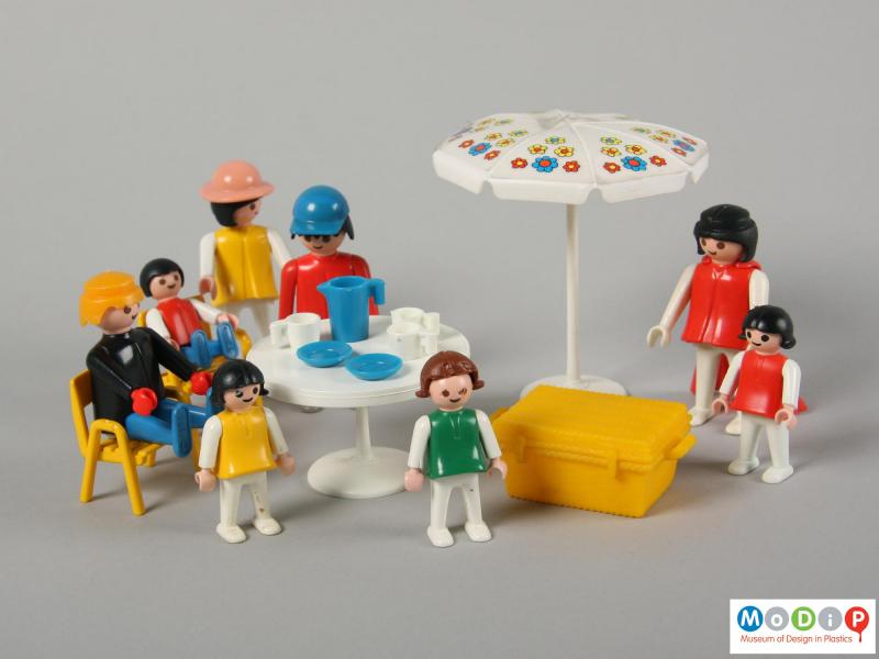 Front view of a picnic scene showing the whole group including the parasol, table, chiars, jug, cups, plates, picnic basket, and all the people.