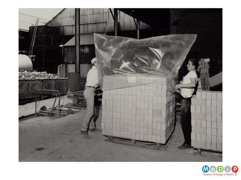 Scanned image showing 2 men covering a pallet load of bricks with a plastic bag.
