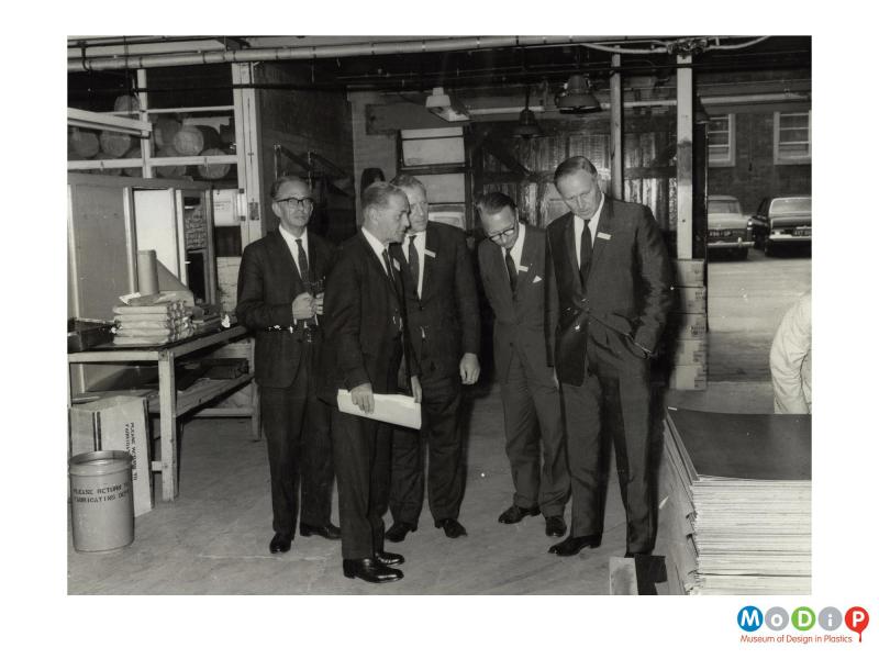 Scanned image showing a group of men wearing suits and looking at a stack of sheet material.