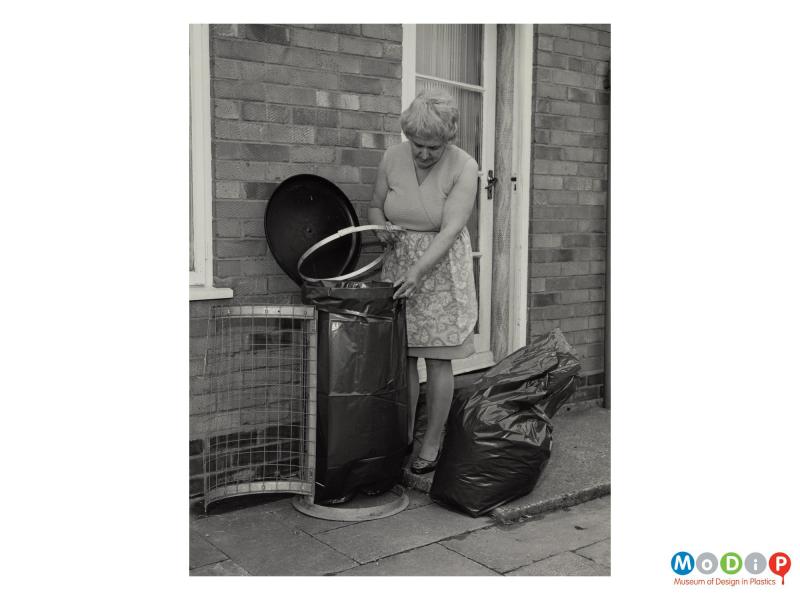 Scanned image showing a woman wearing an apron changing a bin bag in an external bin.