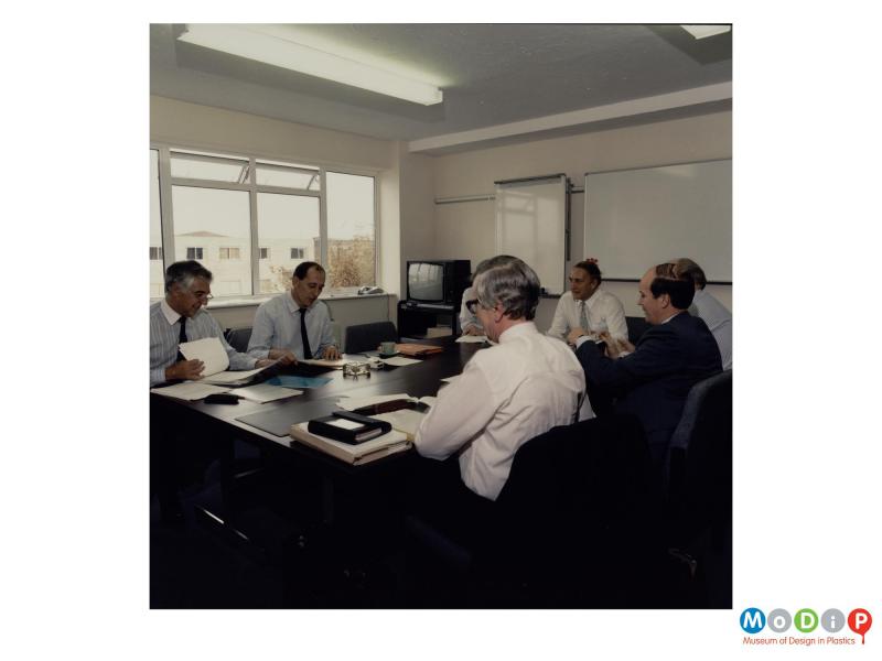 Scanned image showing a group of male employees sitting around a meeting table. 
