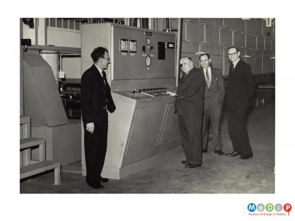 Scanned image showing four men in suits standing at a control panel.