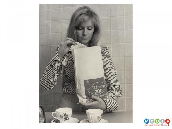 Scanned image showing a lady holding a large plastic bag containing teabags.