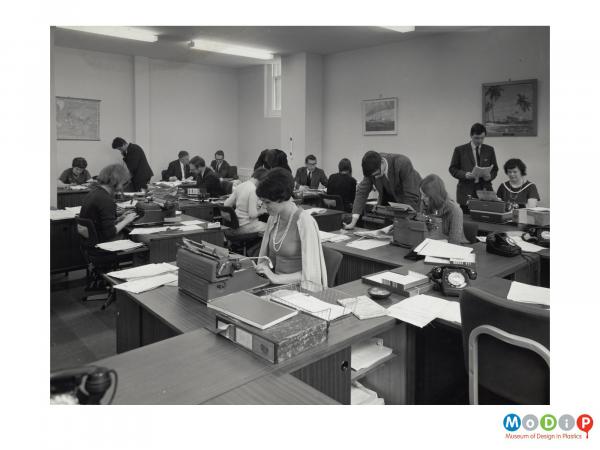 Scanned image showing an office filled with women at typewriters, and suited men.
