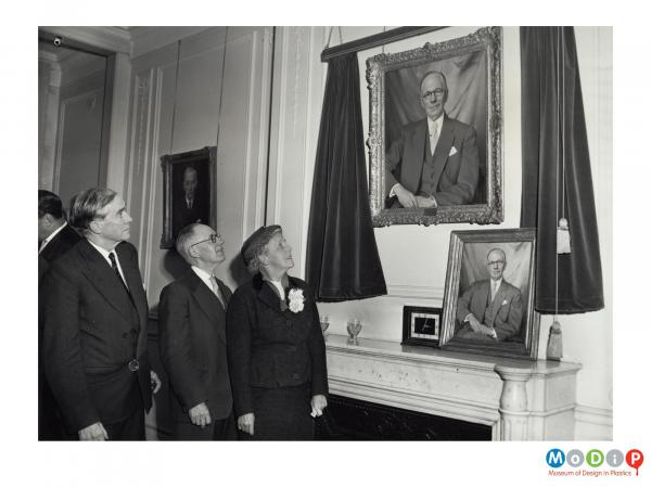 Scanned image showing a group of two men and a woman looking up at a painted portrait of a man.