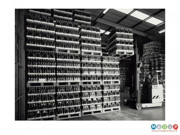 Scanned image showing a man using a forklift truck to move pallet loads of bottles.