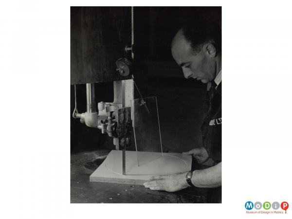 Scanned image showing a man cutting a panel using a mounted bandsaw.
